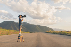 A young boy dressed in paper boy hat and retro attire stands on a stool calling out his message through a megaphone. He is on a rural road in the mountains of Utah County, Utah, USA.